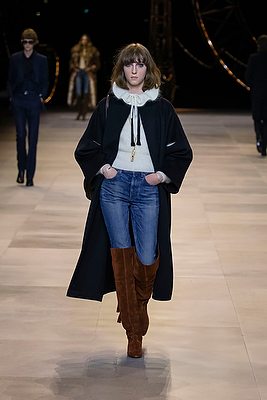 Cette saison, Hedi Slimane imagine une femme au style bourgeois qui forme une déclinaison d'un luxe discret qui séduit immédiatement. Ici un jean porté avec des cuissardes, une chemise lavallière et un élégant manteau façon cape.