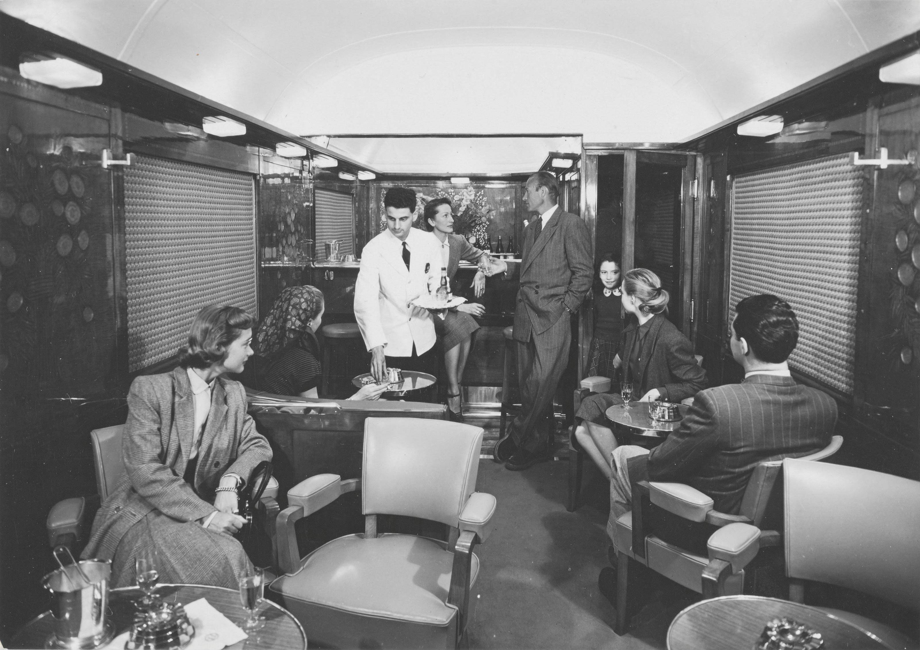 Photographie publicitaire pour la promotion du Train Bleu de la CIWL, reliant paris à la Côte d'Azur, montrant des passagers à bord d'une voiture-salon Pullman transformée en bar, vers 1970. - FDOE - Exposition Orient-Express & Cie