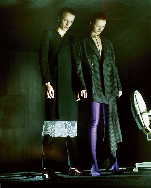 À droite, elle : veste et jupe asymétrique en laine prince-de-galles, chemise en spandex et cuissardes, BALENCIAGA. À gauche, lui : manteau et pantalon en laine, tablier en dentelle et boots, BALENCIAGA.